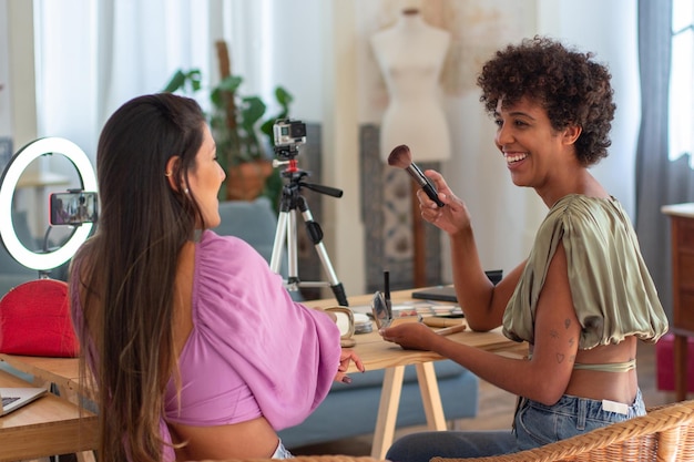 Side view of young female beauty bloggers talking when vlogging. Two women sitting at table demonstrating cosmetic products and explaining how to do make up. Make-up tutorial online, blogging concept