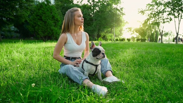 かわいい白と茶色のフレンチブルドッグと新鮮な草の上に座っている若い幸せな女性の側面図。夏の夕日を楽しむゴージャスな笑顔の女の子、都市公園で犬をかわいがります。人間と動物の友情。