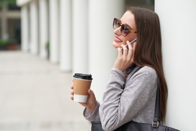 Side view of young lady in sunglasses making phone call leaning on building wall