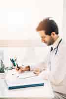 Free photo side view of a young male doctor writing on clipboard in clinic