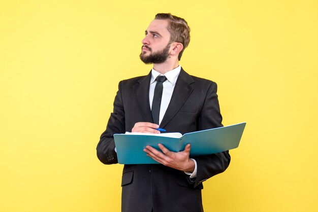 Side view of young man businessman looking up and thinking while pointing pen to the blue folder on the yellow wall