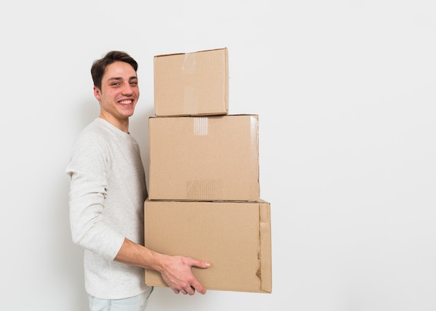 Side view of a young man carrying the stack of cardboard boxes against white wall