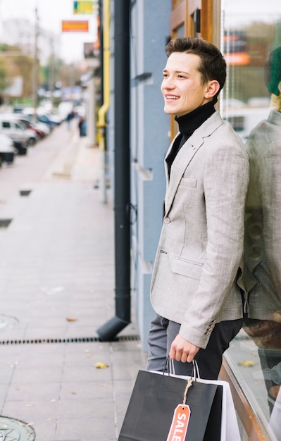 Side view of a young man in fashionable suit holding shopping bags standing on street