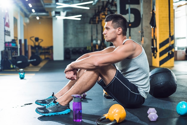 Free photo side view of a young man sitting on floor near exercise equipments and water bottle