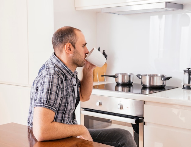 Side view of a young man sitting in the kitchen drinking the coffee