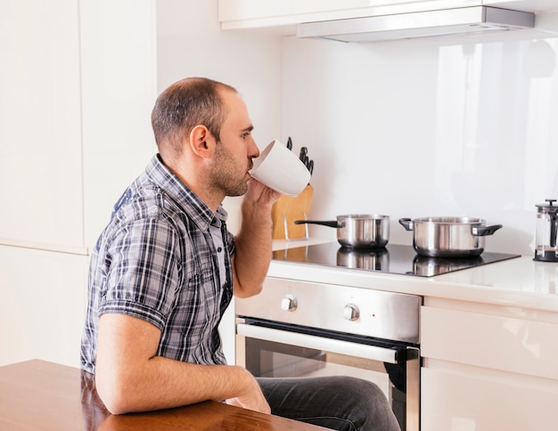Вид сбоку молодого человека, сидящего на кухне и пьющего кофе