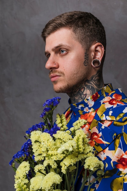 Side view of a young man with pierced ears and nose holding yellow and blue limonium flower against gray background