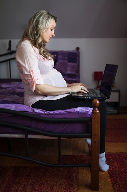 Free photo side view of a young pregnant woman sitting on bed using laptop