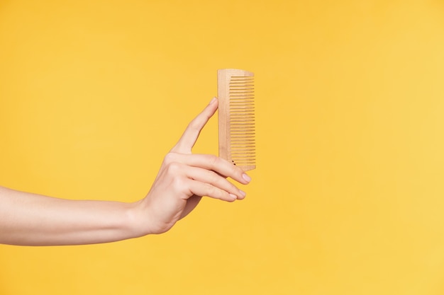 Side view of young well-groomed woman's hands keeping upright wooden while going to comb hair, isolated over orange background. haircare and human hands concept