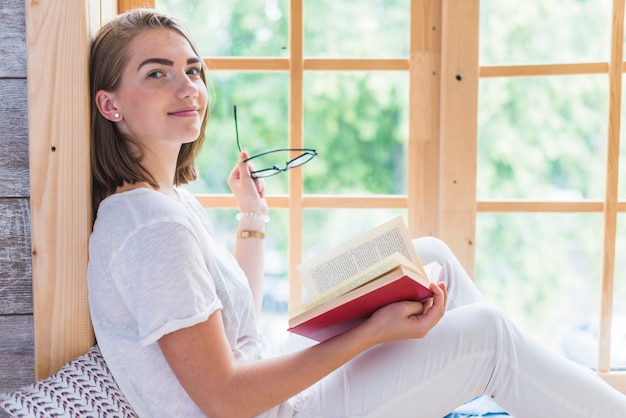 Side view of young woman holding eyeglasses and book near the window