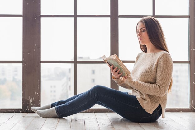 Side view of a young woman sitting near the window reading book