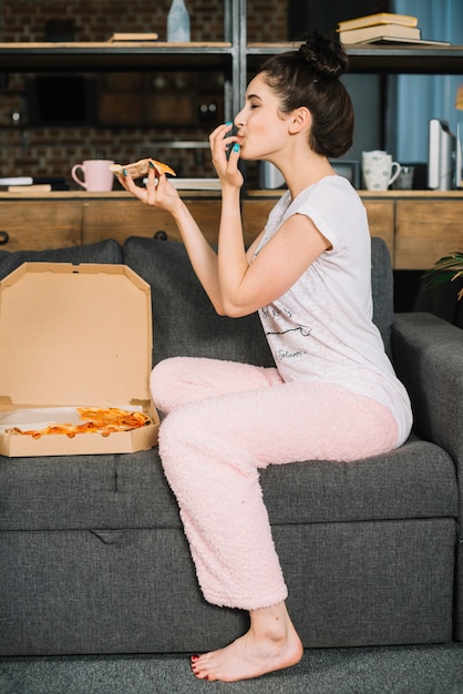 Side view of a young woman sitting on sofa eating pizza