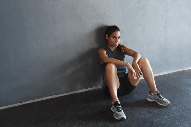 Sideangle shot sassy goodlooking motivated athletic strong sportswoman in activewear taking rest after workout lean on gym wall sit floor waiting fitness instructor start functional training