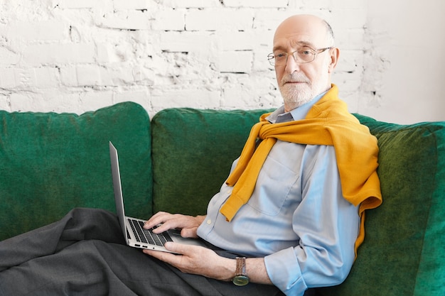 Боковой горизонтальный портрет шестидесятилетнего бородатого мужчины-предпринимателя в очках и свитере над синей рубашкой, работающего удаленно, сидящего на диване с электронным устройством на коленях и смотрящего в камеру