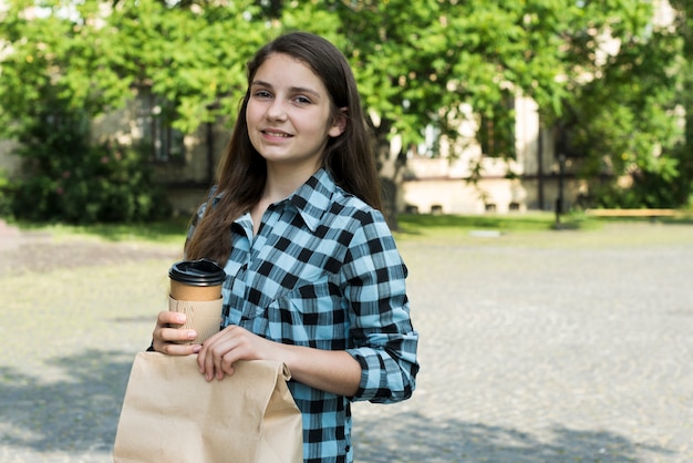 Sideways medium shot of teenage girl holding papercup and lunch bag