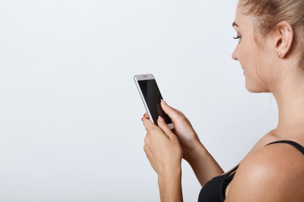 Sideways portrait of female with healthy pure skin holding mobile phone in hands with blank screen, reading news online while using free internet connection. People, modern technologies, communication