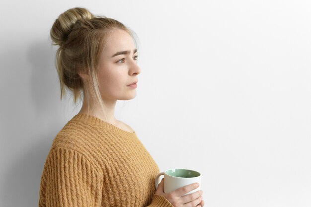 Sideways serious thoughtful young woman with messy hairdo holding large mug, drinking hot cocoa at home, standing at blank wall