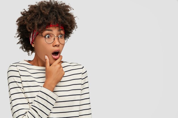 Sideways shot of amazed dark skinned young Afro woman holds hand on chin