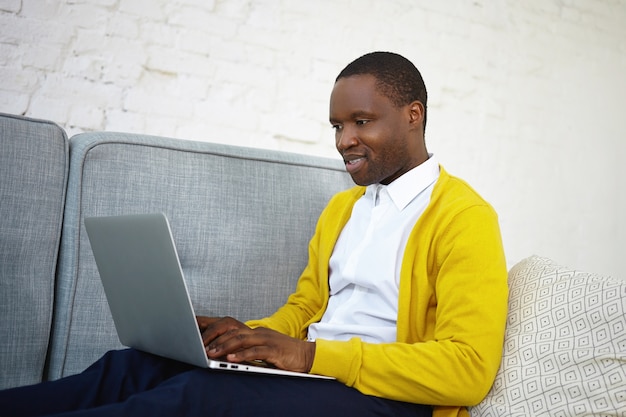 Sideways shot of attractive talented dark skinned male copywriter in stylish wear sitting on couch at home with portable computer on his lap, keyboarding new article for online internet magazine