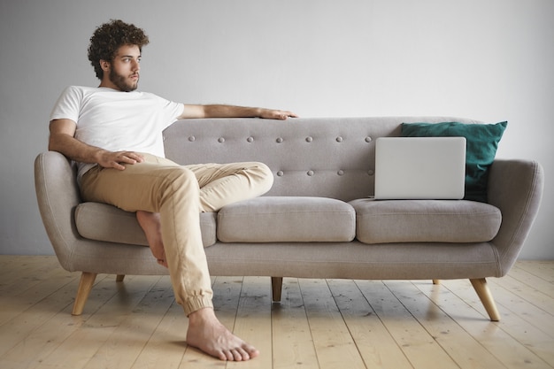 Sideways shot of attractive young Caucasian male with thick beard using portable computer for work, browsing internet, reading articles and surfing websites, sitting comfortably on gray couch