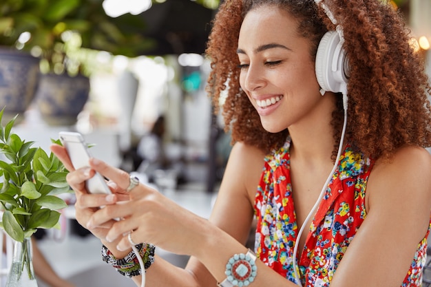 Снимок сбоку: красивая молодая афроамериканка проверяет электронную почту на смартфоне, слушает классную музыку из плейлиста, использует современные наушники