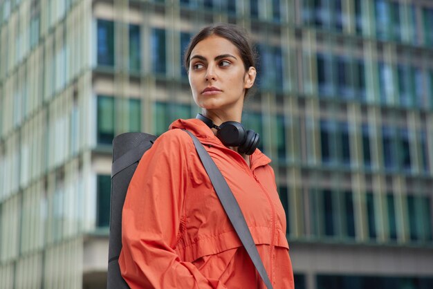 Sideways shot of brunette sportive woman in windbreaker carries rolled kareamt going to have fitness training with instructor poses outdoors