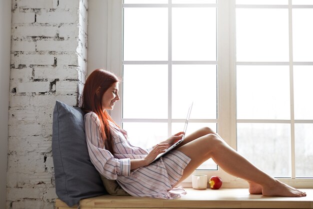 Sideways shot of cheerful teenage girl in striped shirt relaxing by window, speaking to friend via online video chat, using laptop. Pretty woman sitting barefooted on windowsill with portable computer