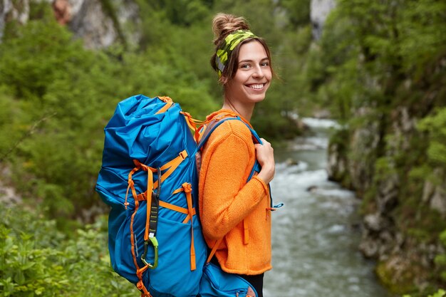 Sideways shot of cheerful woman in orange jumper, wanders near mountain river