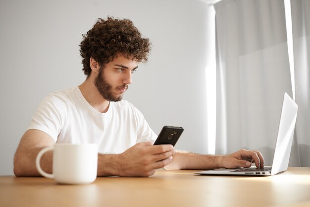 Снимок сбоку: привлекательный молодой бородатый бизнесмен в белой футболке, использующий ноутбук и мобильный для удаленной работы, пьющий утренний кофе, сидящий за деревянным столом с электронными устройствами
