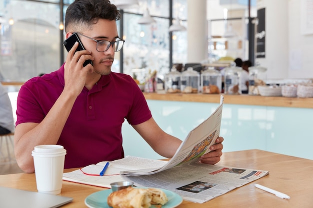 Sideways shot of concentrated entrepreneur reads business newspaper, makes phone call, works remotely from coffee shop, talks on cellular, drinks takeaway coffee, eats dessert. Mass media concept