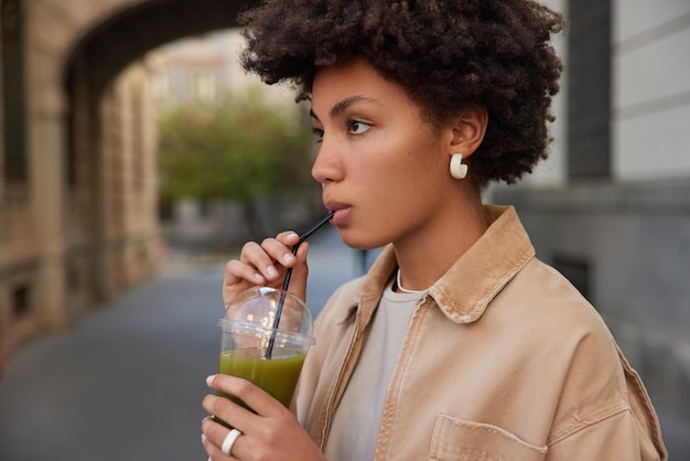 Sideways shot of curly haired woma drinks fresh detox smoothie has healthy nutrition keeps to diet wears brown jacket strolls outdoors against blurred background focused forward Vegetarian meal