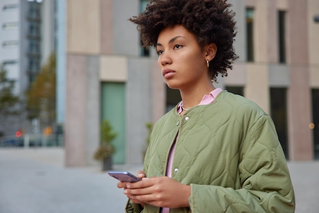 Sideways shot of curly haired young woman surfs mobile phone at street wears streetwear focused pensively