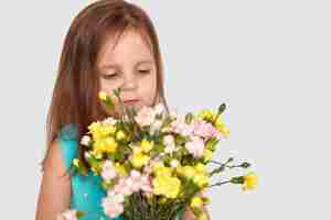 Free photo sideways shot of cute small girl with long hair, enjoys pleasant odour from flowers, dressed in fashionable blue dress, isolated on white with copy space for your promotion or text