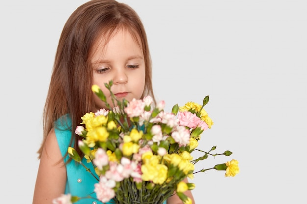 Sideways shot of cute small girl with long hair, enjoys pleasant odour from flowers, dressed in fashionable blue dress, isolated on white with copy space for your promotion or text