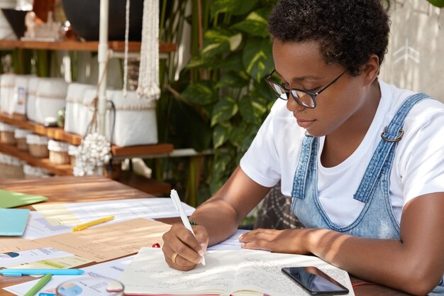 Sideways shot of dark skinned lady in casual clothes, writes down in notebook