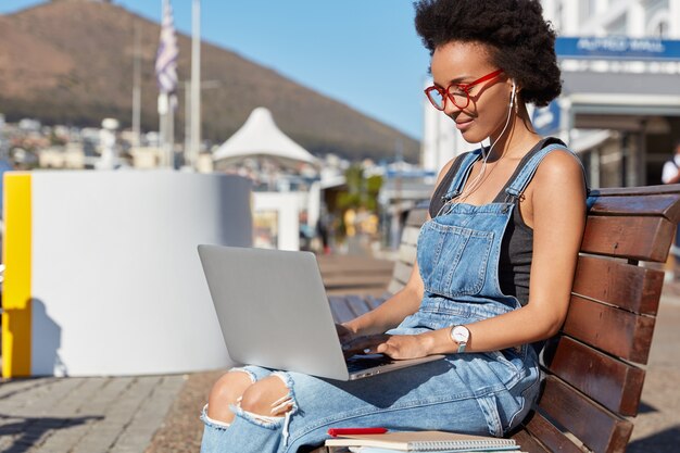 Sideways shot of dark skinned woman with Afro haircut, watches video on laptop computer, holds device on knees, sits at bench in fresh air, studies outdoor,