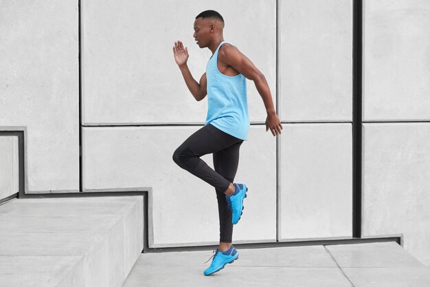 Sideways shot of determined Afro American sportsman runs up stairs, has goal to overcome breath shortage, wears vest and sneakers, poses over white wall. Athlete sporty young man jumps at steps