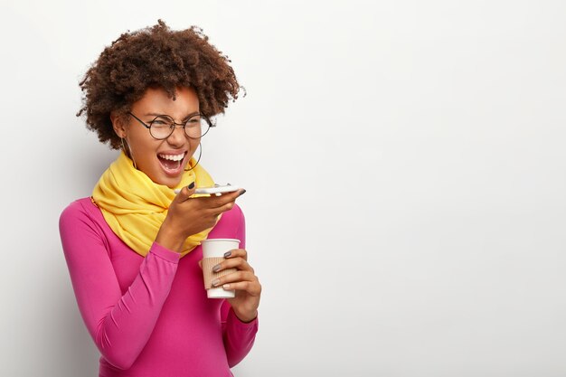 Снимок сбоку эмоциональной темнокожей женщины с вьющейся прической, использует приложение для распознавания голоса на современном мобильном телефоне, держит кофе на вынос, носит очки, розовую водолазку, позирует над белой стеной