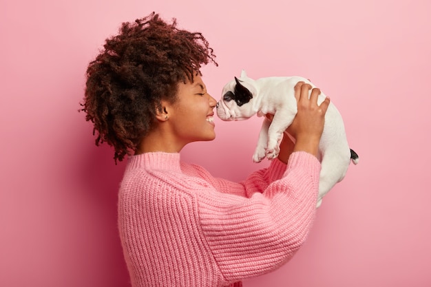 Sideways shot of glad Afro American dog hostess touches noses with small cute puppy