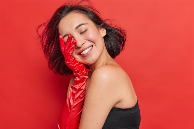 Sideways shot of happy Asian woman keeps eyes closed smiles tenderly covers face with hand wears long elegant gloves and dress shows bare shoulders has healthy clean skin isolated over red studio wall