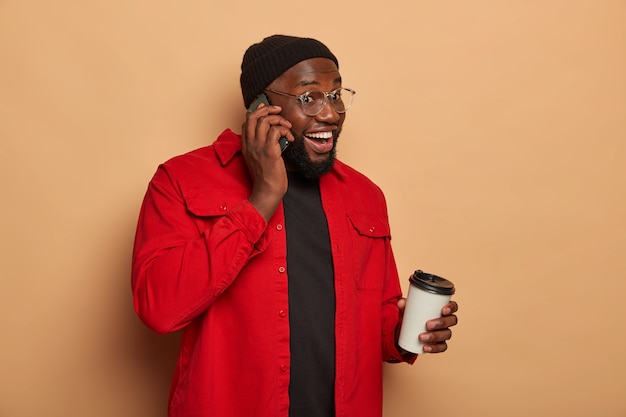 Sideways shot of happy dark skinned guy makes telephone call, drinks takeaway coffee