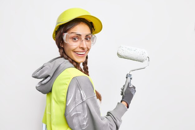 Sideways shot of happy female construction worker smiles gladfully being busy painting 