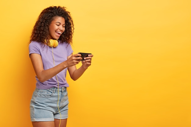 Боковой снимок счастливой дамы с вьющимися волосами, держащей мобильный телефон, смотрящей онлайн-забавный фильм, с позитивной улыбкой на лице