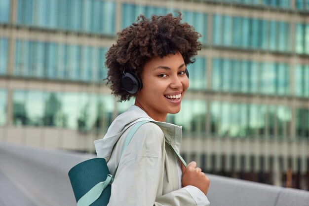 Sideways shot of happy sportswoman poses with fitness mat dressed in anorak listens music via headphones poses against blurred building background. Glad healthy female model prepares for yoga