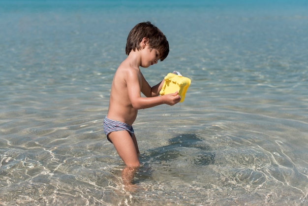 Colpo laterale del bambino che gioca con il secchio di sabbia in acqua