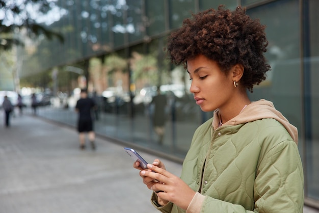 巻き毛のミレニアル世代の女性の横向きのショットは、ソーシャルネットワークでブログを楽しんでいますぼやけた背景タイプに対して都市の屋外でポーズをとるテキストメッセージは携帯電話でニュースを読む新しいアプリを閲覧する