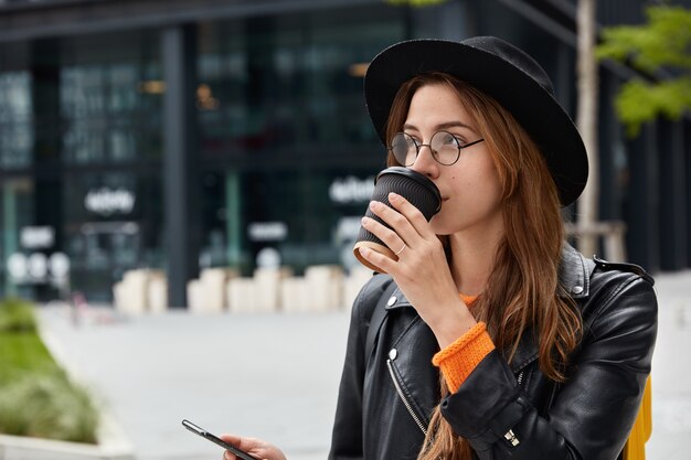 Sideways shot of pensive young girl has coffee break after strolling across city, holds smart phone device, checks email box, focused into distance