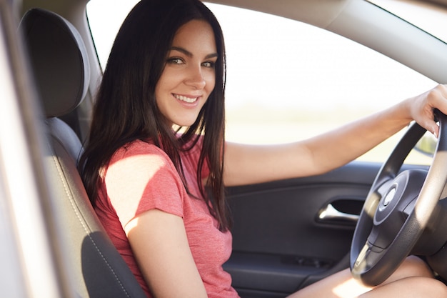 Sideways shot of pleasant looking brunette woman sits in her own automobile instructor