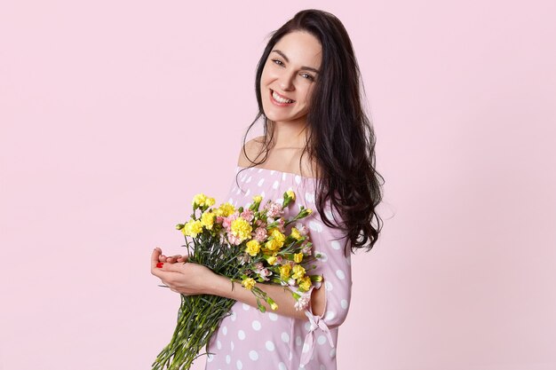 満足して魅力的な若い女性の横向きのショットは、水玉のドレスを着た春の花を保持し、ピンクのポーズ、顔に優しい笑顔、夫とのロマンチックな関係