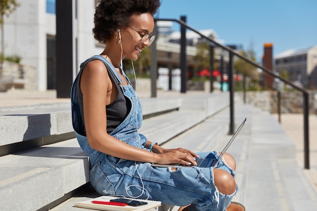 Sideways shot of relaxed carefree young lady with crisp hair, listens radio in earphones, keyboards on laptop computer, does freelance work, diary near, sits at steps during sunny day over city view